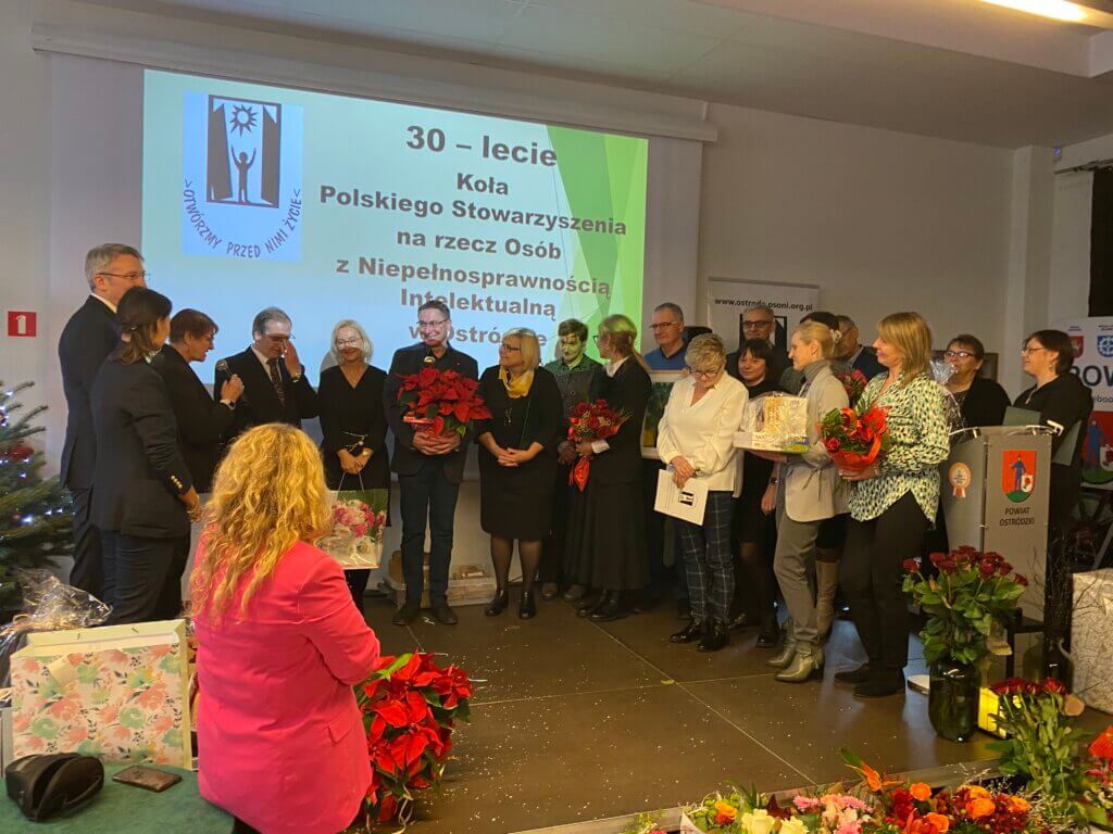 Kilkanaście osób stoi, za nimi na ekranie wyświetlony napis 30-lecie Koła Polskiego Stowarzyszenia na rzecz Osób z Niepełnosprawnością Intelektualną
